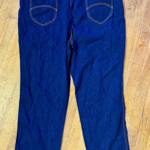 May include: Dark blue denim jeans with a high waist and elastic waistband. The jeans have two back pockets with decorative stitching and a small brand label. The seams are highlighted with tan stitching.