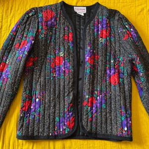 May include: Black quilted jacket with a floral pattern in red, purple, and green. The jacket has a V-neck and long sleeves. The background is a solid yellow color.
