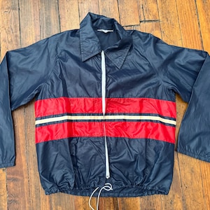 May include: Navy blue jacket with a white zipper and collar. The jacket features a red and white stripe design across the chest. The jacket is made of a lightweight material.