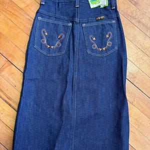 May include: A vintage denim skirt in a dark blue wash. The skirt features decorative stitching and embroidered details on the back pockets. A Wrangler tag is visible. The skirt is a classic, timeless piece.