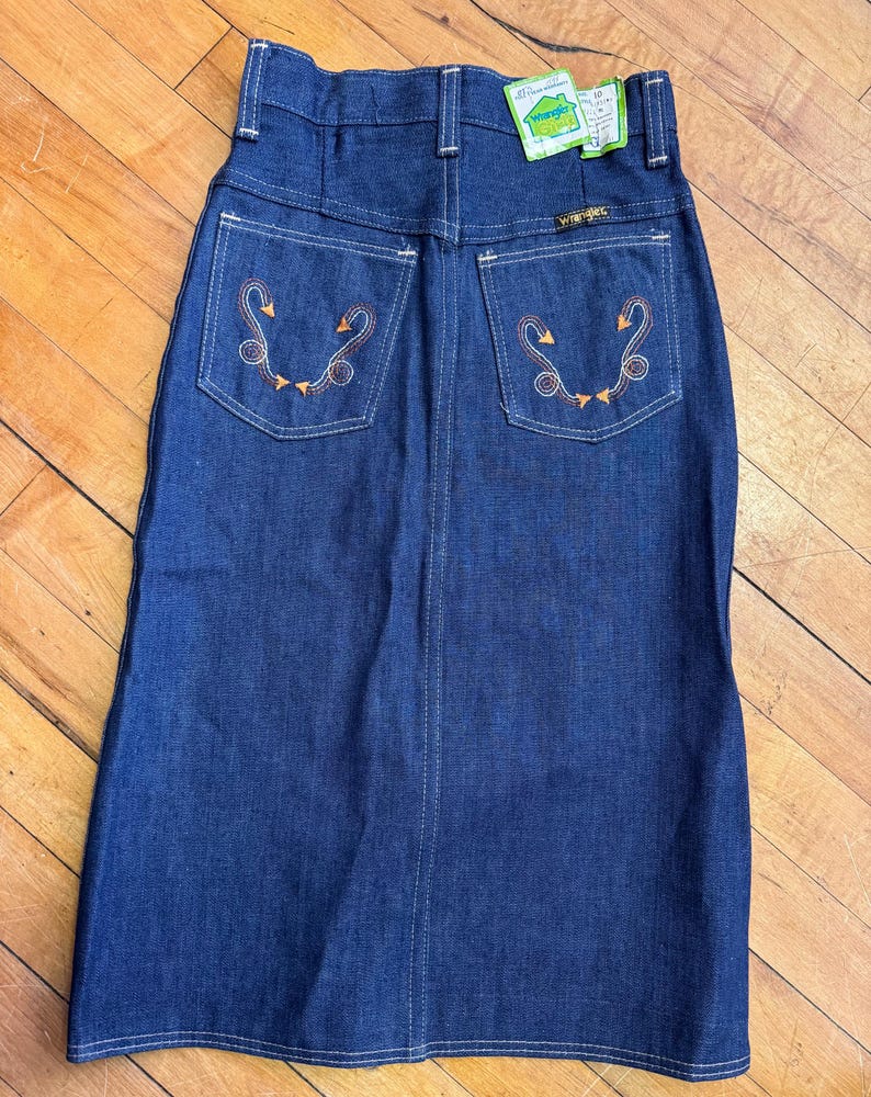 May include: A vintage denim skirt in a dark blue wash. The skirt features decorative stitching and embroidered details on the back pockets. A Wrangler tag is visible. The skirt is a classic, timeless piece.