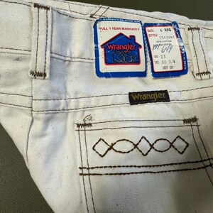 K&ouml;nnte beinhalten: Wei&szlig;e Wrangler Kids Jeans mit braunen N&auml;hten und einer Ges&auml;&szlig;tasche mit einem Rautenmuster. Die Jeans haben ein Etikett mit der Aufschrift "Full 1 Year Warranty" und "Wrangler Kids". Das Etikett enth&auml;lt auch die Gr&ouml;&szlig;e, den Stil und die Losnummer. Die Jeans haben einen braunen Leder-Patch mit der Aufschrift "Wrangler".