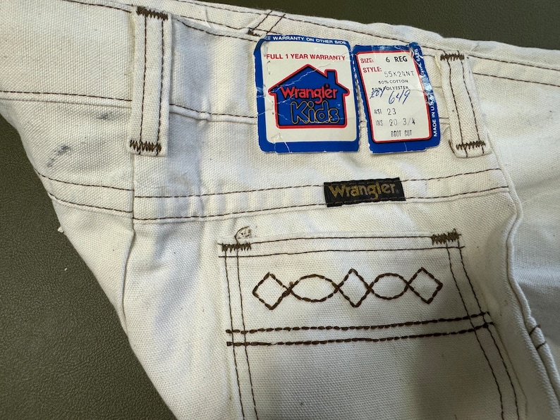 K&ouml;nnte beinhalten: Wei&szlig;e Wrangler Kids Jeans mit braunen N&auml;hten und einer Ges&auml;&szlig;tasche mit einem Rautenmuster. Die Jeans haben ein Etikett mit der Aufschrift "Full 1 Year Warranty" und "Wrangler Kids". Das Etikett enth&auml;lt auch die Gr&ouml;&szlig;e, den Stil und die Losnummer. Die Jeans haben einen braunen Leder-Patch mit der Aufschrift "Wrangler".