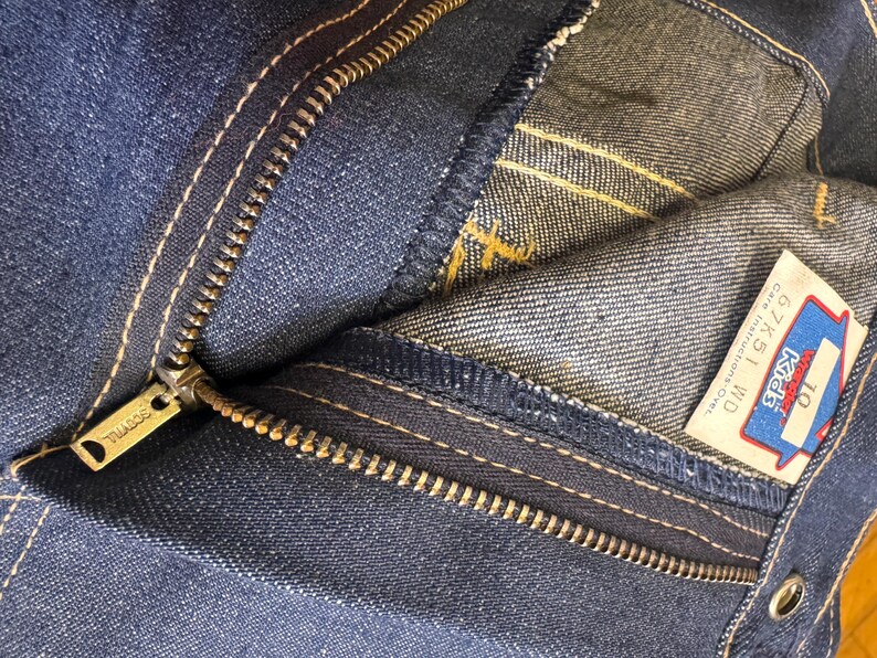 May include: Close-up of blue denim jeans with a visible zipper and white stitching. The zipper pull is gold-colored and has the brand name "SCOVILL" engraved. A tag with the text "Kids" and size "10" is visible.