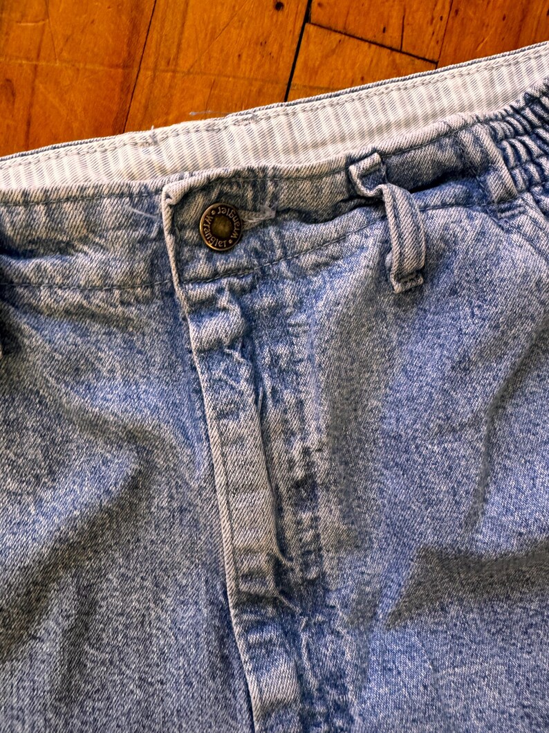 May include: Close-up of light blue denim shorts. The shorts have a button closure with the text "ORIGINAL" on the button. The waistband is elasticized with a striped pattern. The denim has a faded, worn appearance.