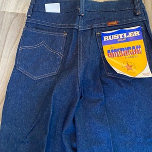 May include: A pair of dark blue denim jeans with a back pocket featuring a Rustler brand label that reads "An American Tradition Crafted with Pride in USA".