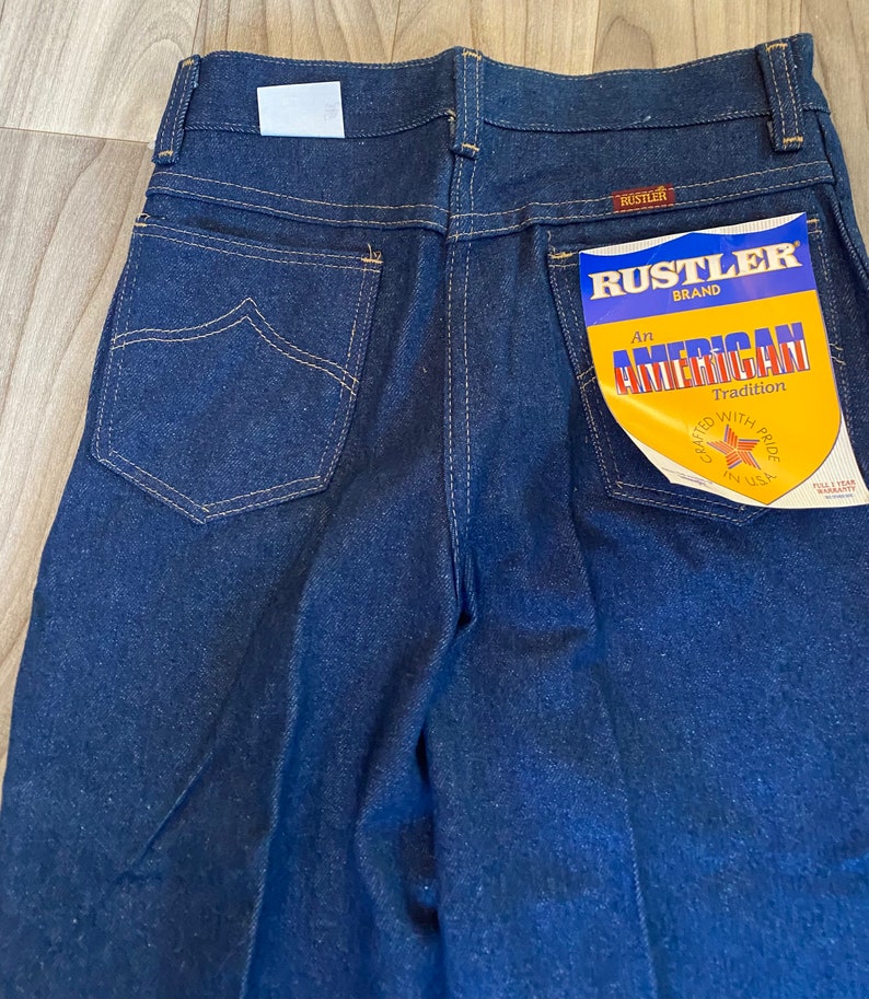 May include: A pair of dark blue denim jeans with a back pocket featuring a Rustler brand label that reads "An American Tradition Crafted with Pride in USA".