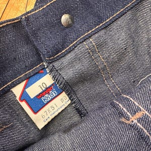 May include: Close-up of blue denim Wrangler Kids jeans. The jeans have a button closure and a tag that reads "10" and "Wrangler Kids". The tag is red, white, and blue. The jeans are size 10.