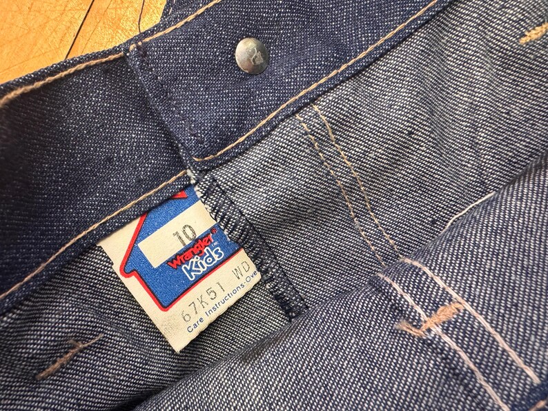 May include: Close-up of blue denim Wrangler Kids jeans. The jeans have a button closure and a tag that reads "10" and "Wrangler Kids". The tag is red, white, and blue. The jeans are size 10.