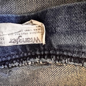 May include: Close-up of a section of blue denim jeans. A white tag with the brand name "Wrangler" is visible, along with size information: 33 x 34. The jeans have a classic, worn look.