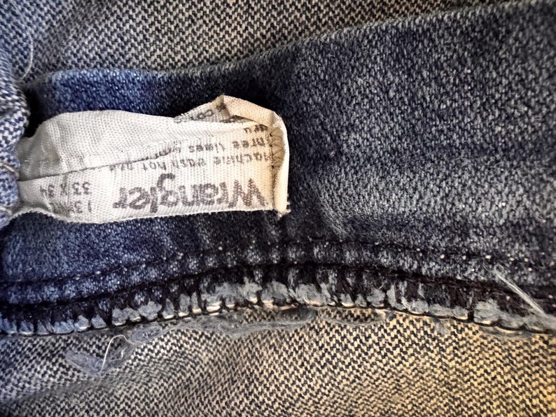 May include: Close-up of a section of blue denim jeans. A white tag with the brand name "Wrangler" is visible, along with size information: 33 x 34. The jeans have a classic, worn look.