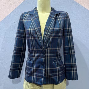 May include: A blue plaid blazer with a notched collar and two front pockets. The blazer has a single-button closure and is displayed on a mannequin. The plaid pattern includes white, red, and tan lines.