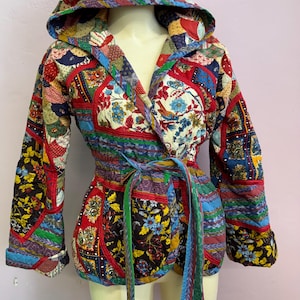 May include: A hooded jacket constructed from a patchwork of quilted fabric featuring various floral, geometric, and abstract patterns in red, blue, yellow, and black. The jacket includes long sleeves, a wrap-style front, and a matching fabric tie belt.