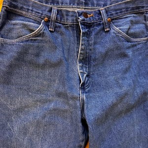 May include: Close-up of blue denim jeans. The jeans have a button and zipper closure, front pockets, and belt loops. The denim has a faded appearance, with subtle variations in color. The jeans are a classic style.
