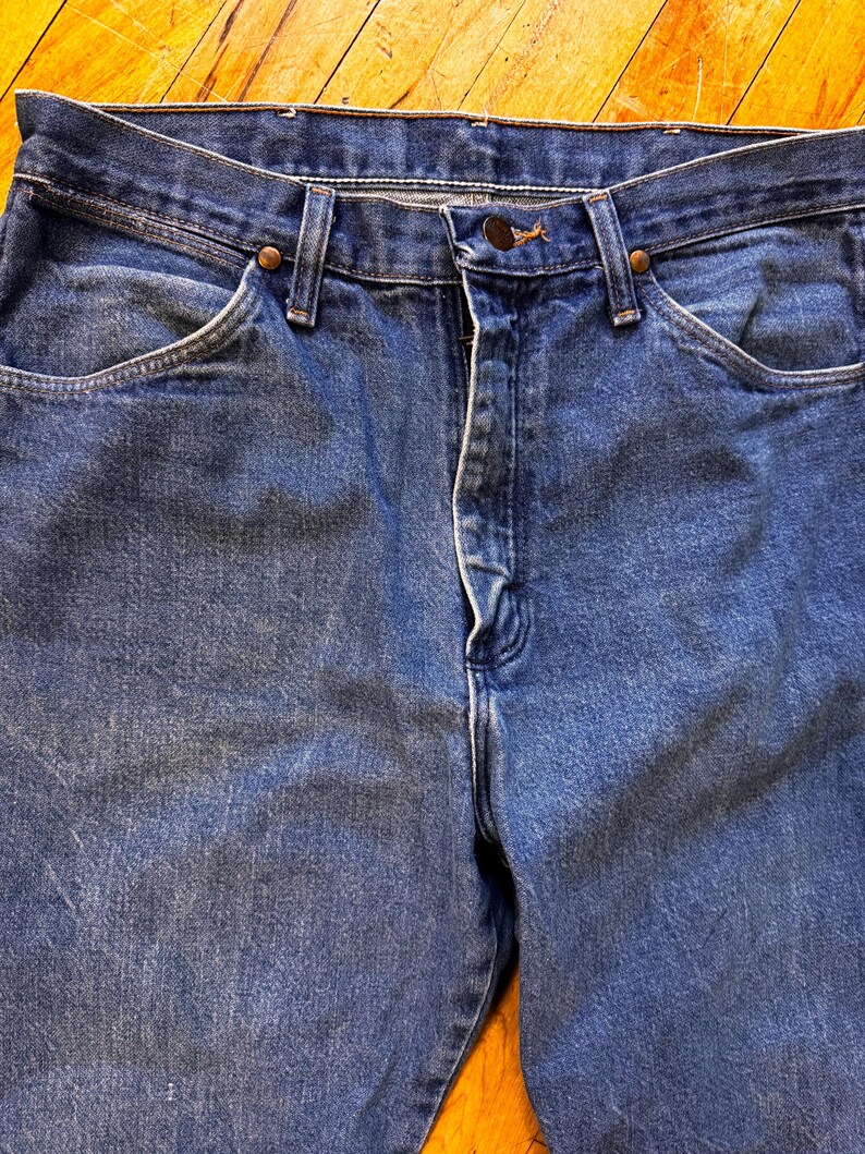 May include: Close-up of blue denim jeans. The jeans have a button and zipper closure, front pockets, and belt loops. The denim has a faded appearance, with subtle variations in color. The jeans are a classic style.