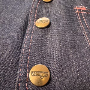 Może przedstawiać: Zbliżenie na ciemnoniebieski dżinsowy element garderoby z trzema mosiężnymi guzikami. Każdy guzik jest wytłoczony nazwą marki "Wrangler". R&oacute;żowe przeszycia podkreślają szwy i kieszeń.