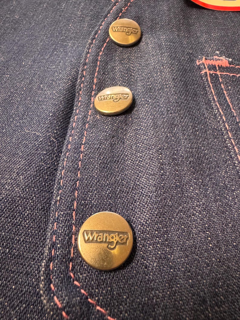 Może przedstawiać: Zbliżenie na ciemnoniebieski dżinsowy element garderoby z trzema mosiężnymi guzikami. Każdy guzik jest wytłoczony nazwą marki "Wrangler". R&oacute;żowe przeszycia podkreślają szwy i kieszeń.