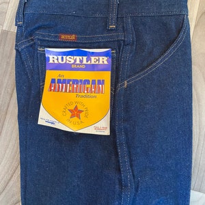 May include: A pair of dark blue denim jeans with a back pocket and a yellow and blue label that reads "Rustler Brand An American Tradition Crafted with Pride in U.S.A. Full 1 Year Warranty".