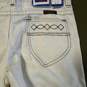 K&ouml;nnte beinhalten: Wei&szlig;e Wrangler Kids Jeans mit braunen N&auml;hten und einem Diamantenmuster auf der Ges&auml;&szlig;tasche. Die Jeans haben eine Bootcut-Form und sind in Gr&ouml;&szlig;e 6 Regular. Das Etikett zeigt 65% Baumwolle und 35% Polyester.