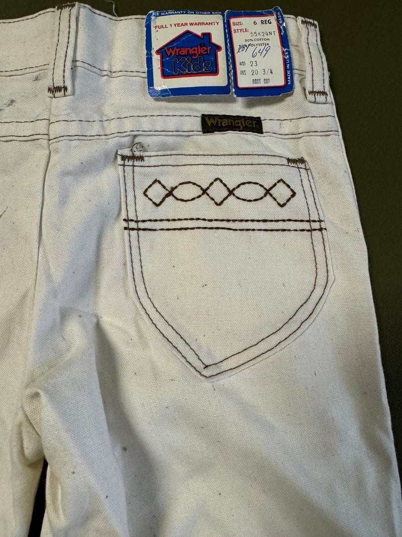 K&ouml;nnte beinhalten: Wei&szlig;e Wrangler Kids Jeans mit braunen N&auml;hten und einem Diamantenmuster auf der Ges&auml;&szlig;tasche. Die Jeans haben eine Bootcut-Form und sind in Gr&ouml;&szlig;e 6 Regular. Das Etikett zeigt 65% Baumwolle und 35% Polyester.