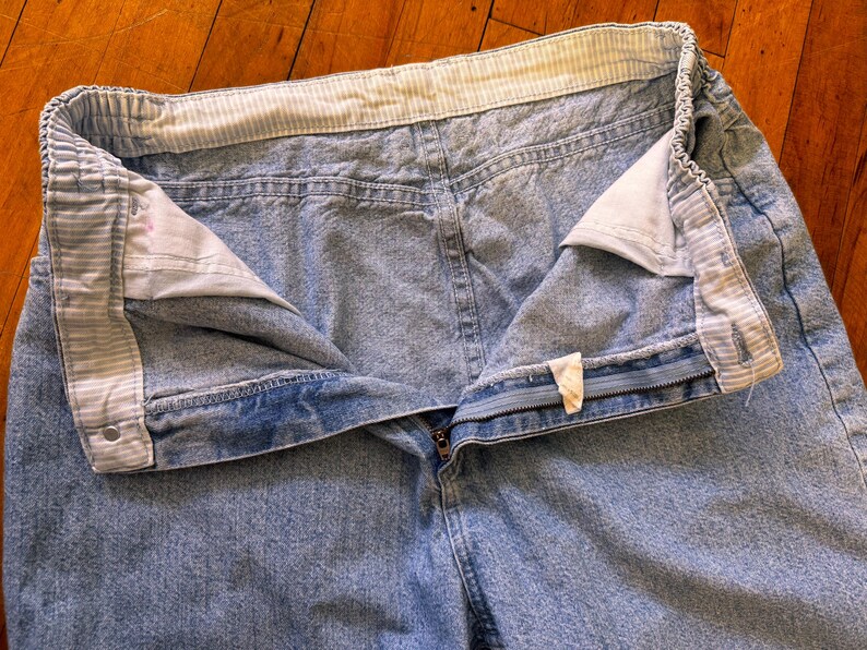May include: Light blue denim jeans with an elastic waistband and a zipper fly. The waistband has a striped pattern. The jeans are unbuttoned, showing the interior lining and a small tag.