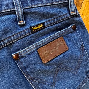 May include: Close-up of blue denim jeans with a back pocket. The pocket features a brown leather patch with the brand name "Wrangler" stitched in a gold color. The jeans have a classic design with visible stitching and a small brand label.