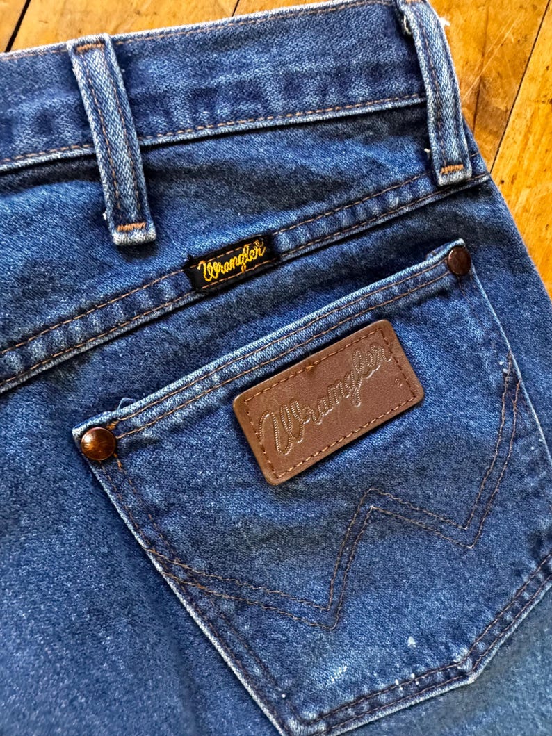 May include: Close-up of blue denim jeans with a back pocket. The pocket features a brown leather patch with the brand name "Wrangler" stitched in a gold color. The jeans have a classic design with visible stitching and a small brand label.