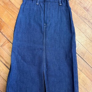May include: A dark blue denim skirt with a front slit. The skirt has a high waist, belt loops, and a button closure. White stitching accents the seams and the front zipper. The skirt is made of a sturdy denim material.