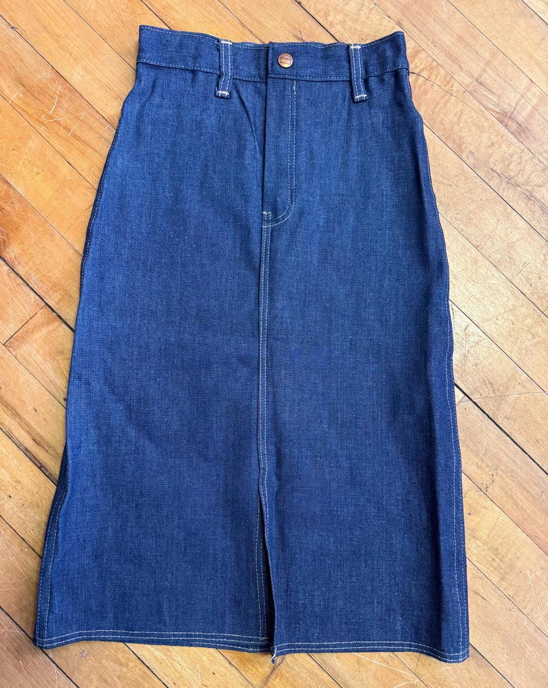 May include: A dark blue denim skirt with a front slit. The skirt has a high waist, belt loops, and a button closure. White stitching accents the seams and the front zipper. The skirt is made of a sturdy denim material.