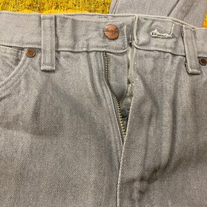 Puede incluir: Un par de pantalones vaqueros grises con cierre de cremallera y bot&oacute;n. Los vaqueros tienen una etiqueta de la marca Wrangler en la parte posterior de la cintura.