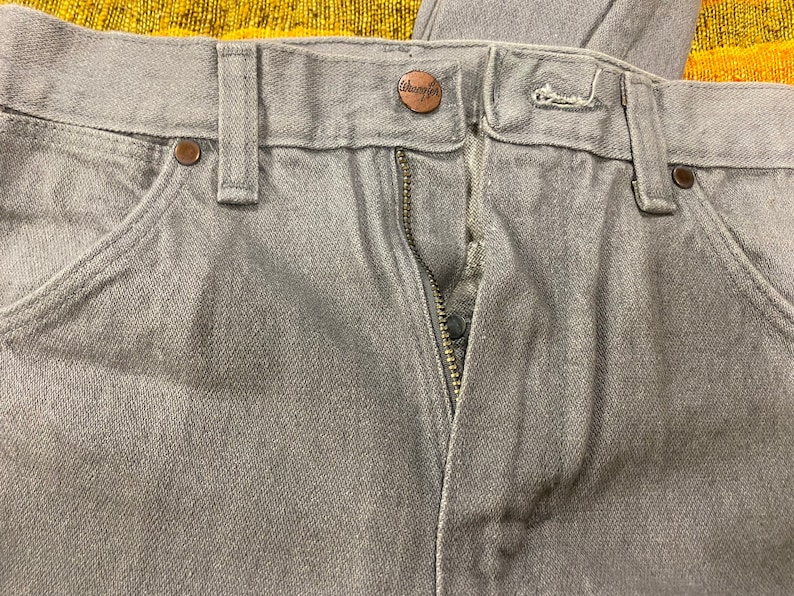 Puede incluir: Un par de pantalones vaqueros grises con cierre de cremallera y bot&oacute;n. Los vaqueros tienen una etiqueta de la marca Wrangler en la parte posterior de la cintura.