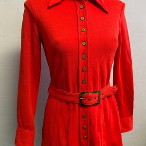 May include: A bright red long-sleeved dress with a collared neckline and a matching belt. The dress features a button-down front with gold-colored buttons and a rectangular belt buckle. The fabric appears to be a soft knit material.
