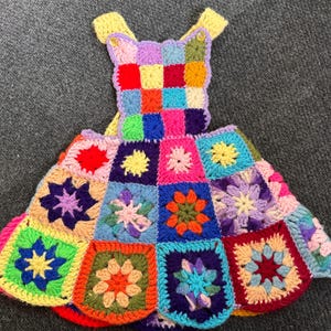 May include: A colorful, handmade crochet dress featuring a patchwork design. The dress is composed of various square motifs in vibrant colors like red, blue, green, and purple, with floral patterns in the center of each square. The dress has yellow straps.