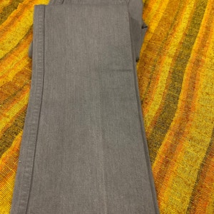 Puede incluir: Un par de pantalones vaqueros de mezclilla gris doblados sobre una tela rayada amarilla y naranja. Los vaqueros tienen un peque&ntilde;o bolsillo en la parte delantera con una etiqueta que dice "Frontier of Change".
