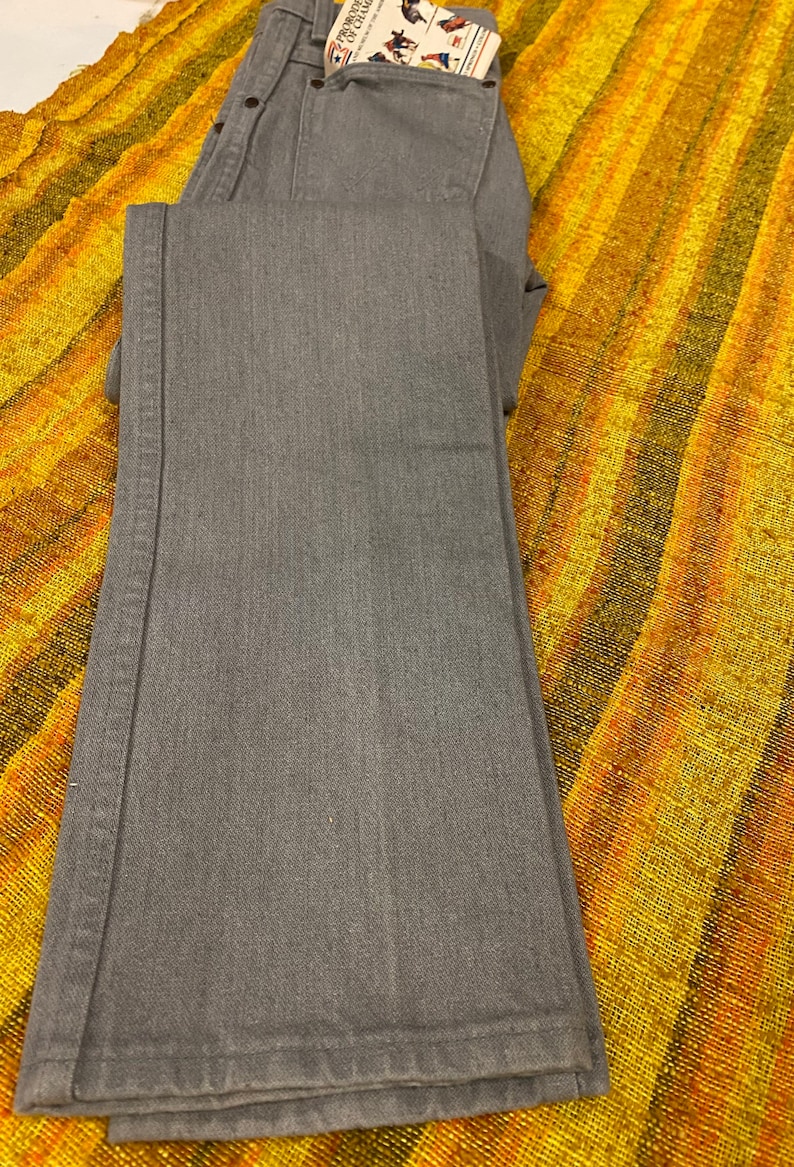 Puede incluir: Un par de pantalones vaqueros de mezclilla gris doblados sobre una tela rayada amarilla y naranja. Los vaqueros tienen un peque&ntilde;o bolsillo en la parte delantera con una etiqueta que dice "Frontier of Change".
