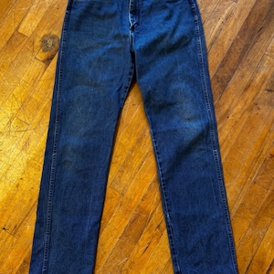 May include: Dark blue denim jeans with a classic straight-leg fit. The jeans feature a button and zipper closure, front and back pockets, and belt loops. The denim has some fading and wear, giving it a vintage look.