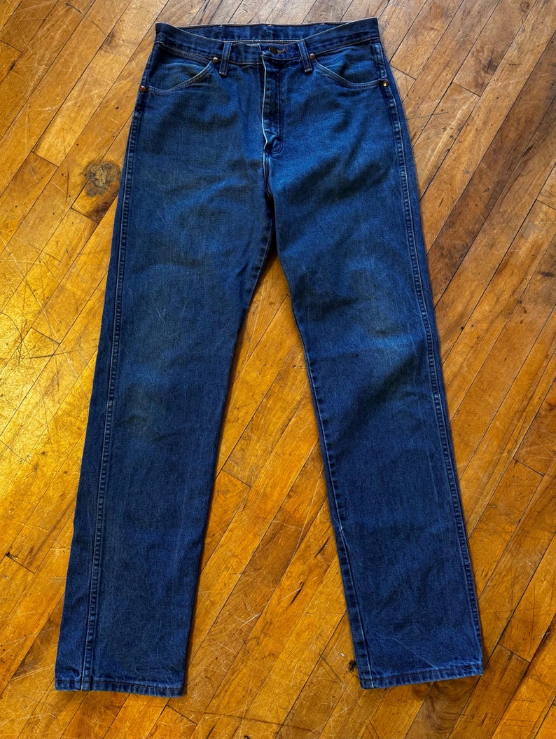 May include: Dark blue denim jeans with a classic straight-leg fit. The jeans feature a button and zipper closure, front and back pockets, and belt loops. The denim has some fading and wear, giving it a vintage look.