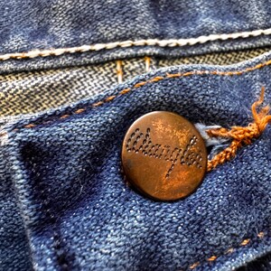 May include: Close-up of blue denim fabric with a copper-colored button. The button is embossed with the word "Wrangler". The denim has white and orange stitching.