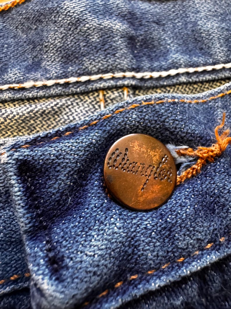 May include: Close-up of blue denim fabric with a copper-colored button. The button is embossed with the word "Wrangler". The denim has white and orange stitching.