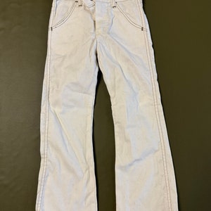 K&ouml;nnte beinhalten: Wei&szlig;e Jeans mit Schlag und braunen N&auml;hten. Die Hose hat einen Knopfverschluss und G&uuml;rtelschlaufen.