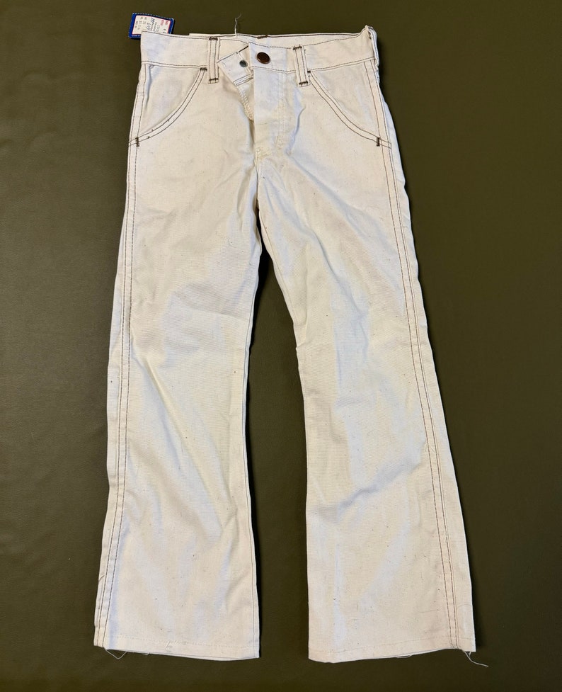 K&ouml;nnte beinhalten: Wei&szlig;e Jeans mit Schlag und braunen N&auml;hten. Die Hose hat einen Knopfverschluss und G&uuml;rtelschlaufen.