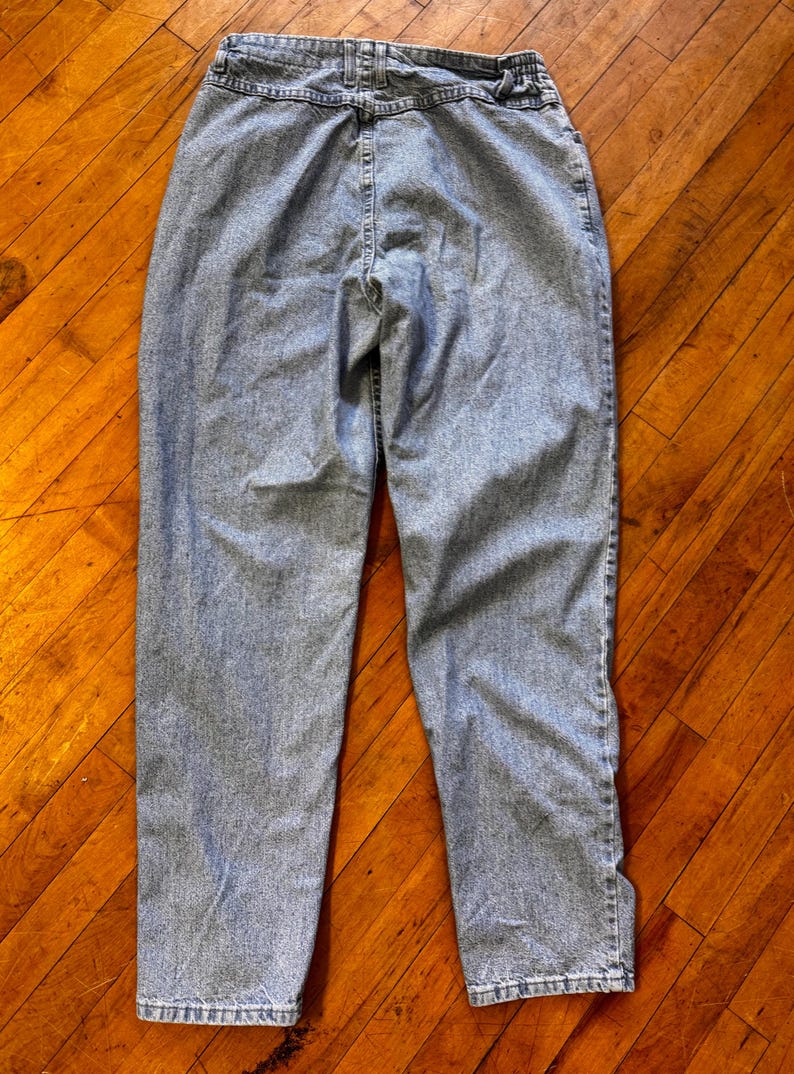 May include: Light blue denim jeans with a high waist and a classic straight leg fit. The jeans have a button and zipper closure, belt loops, and pockets. The back view shows the stitching details and the overall shape of the pants.