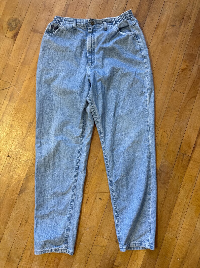 May include: Light blue denim jeans with an elastic waistband and a button closure. The jeans have a classic five-pocket design and a relaxed fit. The denim is a medium wash, and the jeans appear to be vintage.
