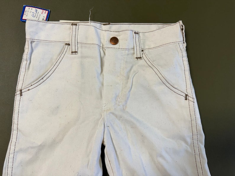 K&ouml;nnte beinhalten: Wei&szlig;e Jeanshose mit braunen N&auml;hten und einem kupferfarbenen Knopf. Die Hose hat einen klassischen F&uuml;nftaschen-Stil.
