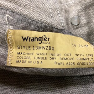 Puede incluir: Un primer plano de una etiqueta de mezclilla Wrangler gris con el n&uacute;mero de estilo 13MWZBG, talla 14 slim, e instrucciones de cuidado. La etiqueta dice "Machine wash inside out, with like colors, tumble dry remove promptly. Made in USA RWPL 6428 6F0S11002".