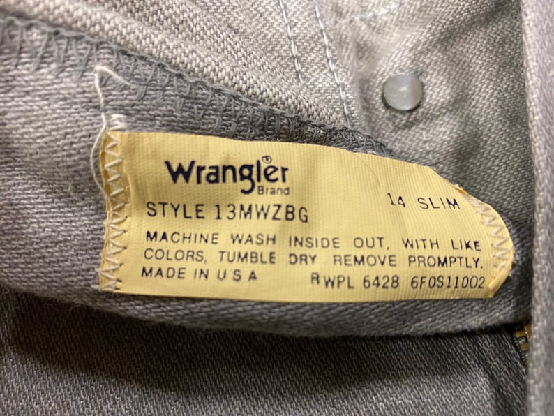 Puede incluir: Un primer plano de una etiqueta de mezclilla Wrangler gris con el n&uacute;mero de estilo 13MWZBG, talla 14 slim, e instrucciones de cuidado. La etiqueta dice "Machine wash inside out, with like colors, tumble dry remove promptly. Made in USA RWPL 6428 6F0S11002".