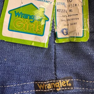 May include: Close-up of a blue denim jean skirt with a Wrangler label and attached tags. The tags read "Wrangler Girls" and include size, style, and material information. The skirt is made in the USA.