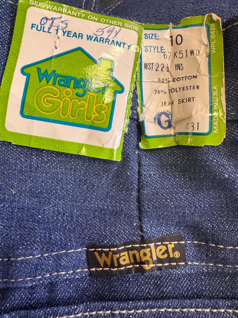 May include: Close-up of a blue denim jean skirt with a Wrangler label and attached tags. The tags read "Wrangler Girls" and include size, style, and material information. The skirt is made in the USA.