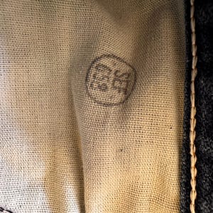 May include: Close-up of a beige fabric with a circular, faded gray stamp. The stamp contains indecipherable characters. The fabric is next to a dark blue denim material with visible stitching.