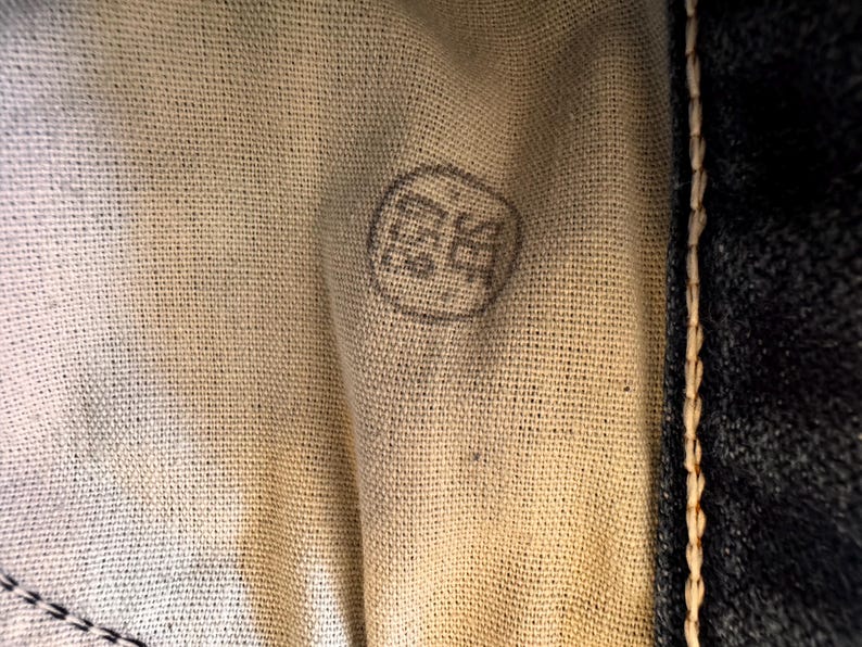 May include: Close-up of a beige fabric with a circular, faded gray stamp. The stamp contains indecipherable characters. The fabric is next to a dark blue denim material with visible stitching.